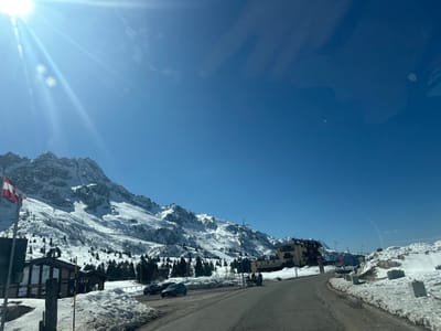 The image presents a snow-covered mountain landscape under a clear blue sky with a bright sun creating lens flare in the upper left quadrant. The scene is located in Vermiglio, Italy. Dominating the background are jagged, snow-capped mountains exhibiting both pristine white slopes and visible rock formations. Coniferous trees are scattered across the lower mountain slopes and in the valleys.

A paved asphalt road, appearing wet in sections, traverses the mid-ground and curves towards the right, leading into the distance. Snow covers the ground extensively, accumulating along the roadside and partially obscuring the landscape.

On the left side of the road, a flagpole displays a Swiss flag (red with a white cross). Behind it, a multi-story building with a dark roof and light-colored walls is partially visible, with a sign indicating "BAR." Two dark-colored passenger vehicles are parked or stopped on the left shoulder of the road near this building, oriented away from the viewer.

Further along the road on the right, a multi-story building with a light yellow/beige facade and a dark roof is situated amidst the snow. Concrete or stone bollards are visible along the right edge of the road, partially buried in snow. No individuals are discernible within the frame, and no specific actions or interactions are depicted.