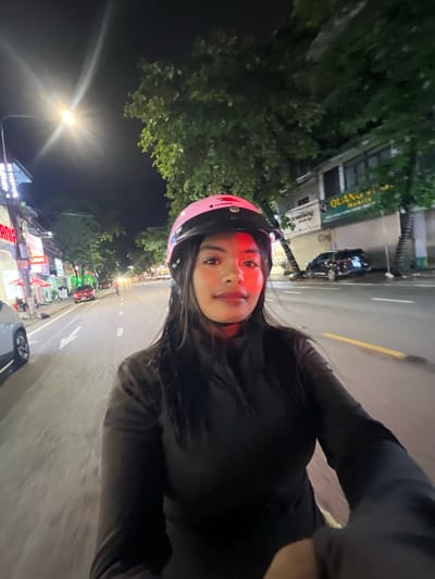 The image is a selfie taken at night from the perspective of someone riding a motorcycle. The subject is a woman wearing a pink helmet and a black jacket. She is looking directly at the camera with a neutral expression. The background shows a street scene with buildings, trees, streetlights, and other vehicles. The street appears to be well-lit, indicating an urban environment. The presence of businesses with signage suggests this is likely a commercial area within Minh Xuân Ward, Vietnam, possibly near a main road given the traffic. The overall atmosphere is that of a typical city night, with the artificial lights illuminating the scene.