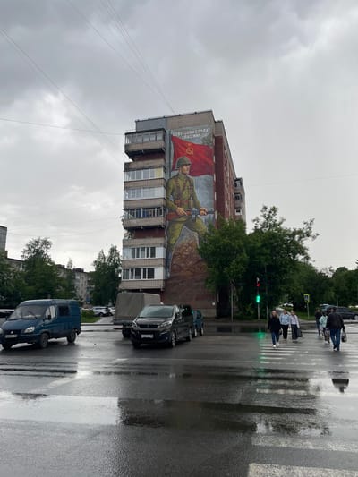 A mural of a Soviet soldier is painted on the side of a multi-story apartment building in Cherepovets, Russia. The soldier, wearing a helmet and holding a rifle, stands in front of a red flag with a hammer and sickle. The mural is located at the intersection of Karl Marx Street and Moskovsky Prospekt. The street is wet from rain, and traffic lights are visible. Several cars and pedestrians are crossing the street. The sky is overcast, and the overall atmosphere is somber.