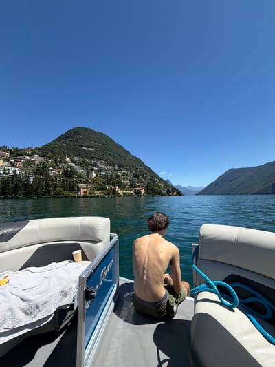 In Lugano, Switzerland, a young person is seated on the rear deck of a boat, facing away from the camera. The person is shirtless, wearing green shorts, and their back is visible, showing a lean physique. The boat, a pontoon style, has white and grey seating and a blue railing with a boat nameplate. A white towel and a paper cup are on the deck to the left of the person. A bright blue rope is coiled on the right side of the boat. The boat is on a body of water, likely Lake Lugano, with a lush green mountain covered in buildings and trees dominating the background on the left side. The sky is clear and blue. To the right, the lake extends towards more mountains in the distance. The scene captures a moment of leisure on the water on a sunny day.