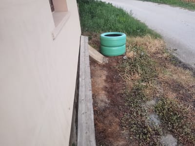 In the city of Lom, Bulgaria, on the outskirts of a building, two green painted tires are stacked on top of each other. They are placed near a wooden plank and a concrete curb. The tires are positioned on the ground, which is covered with dry grass and some green vegetation. A paved road can be seen in the background, bordered by a grassy area. The scene appears to be outdoors, possibly in a residential or semi-rural area.