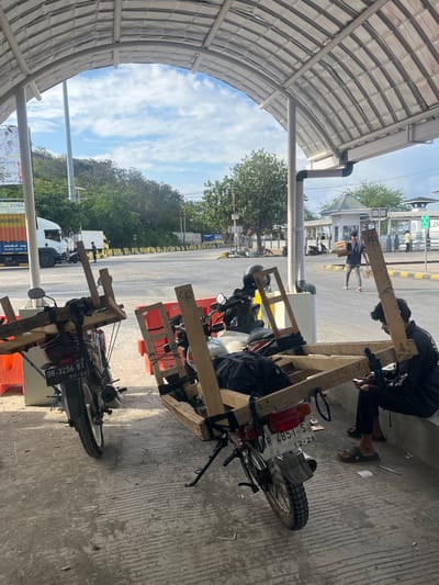 The image depicts a scene at Upt Tambak Sari, Indonesia, under a covered structure. In the foreground, two motorcycles are parked, each fitted with a wooden frame structure. One motorcycle, on the left, has the license plate "DR 3256 BT". The other motorcycle, on the right, has the license plate "DR 4851 S". A person in dark clothing is seated on a ledge to the right of the motorcycles, looking at a mobile phone. In the background, there is a paved area with parked trucks and other vehicles. Several people are visible in the distance, and a small white building with a distinctive roof structure is present. The sky is cloudy. The overall environment appears to be an outdoor waiting or loading area.