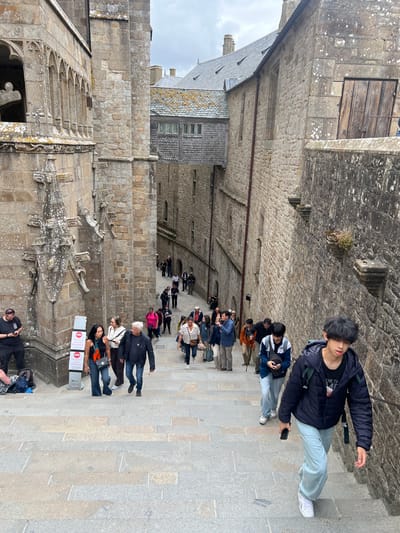 The image shows a group of tourists ascending a wide stone staircase at Le Mont-Saint-Michel, France. The architecture is ancient and made of stone, with arched windows and weathered walls. People are dressed in casual attire, and many are carrying backpacks. The staircase is paved with large, rectangular stones, and there are railings on the right side of the image. The sky is overcast, suggesting a cloudy day. The scene depicts a typical tourist experience at this historical landmark.