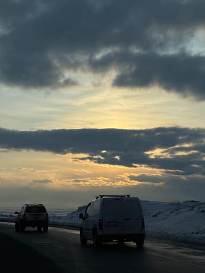 The image presents a late afternoon or early evening road scene in Jraber, Armenia, under a heavily clouded sky with a visible sunset. Two vehicles are observed traveling on a wet, paved two-lane road. In the immediate foreground, a white panel van, fitted with a black roof rack, moves away from the viewer. Its rear license plate is partially discernible as "EW 221 NV". Further along the same road, a dark-colored sport utility vehicle (SUV) also proceeds away from the camera. To the right of the roadway, the terrain is covered in snow, forming gentle, undulating banks. The distant horizon to the left reveals a vast, flat, or gently rolling landscape under a hazy sky. The upper portion of the frame is dominated by dark, dense stratocumulus clouds, while a horizontal band of golden-yellow light from the setting sun is visible between cloud layers near the horizon, casting an orange glow. No individuals are visible.
