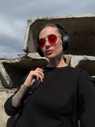This media file features a person wearing black headphones and round red-tinted sunglasses, looking towards the camera. The person has light-colored hair, a tattoo on their neck, and colorful tattoos on their arm. They are wearing a black long-sleeved shirt. Their right hand is holding onto a black strap, likely from a bag or backpack. The scene appears to be outdoors, with a backdrop of grey, cloudy skies and what looks like concrete debris. The image is oriented vertically, with the top of the person's head and headphones at the bottom of the frame. The lighting suggests it might be daytime, possibly overcast. The presence of concrete rubble might indicate a construction site, an abandoned area, or a site damaged by industrial activity, fitting with the context of Norilsk, Russia, a city known for its industrial landscape and often harsh weather conditions. No visible text is present in the image.