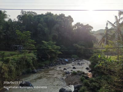 The image depicts a scenic view of a river flowing through a lush, green landscape in Kecamatan Kuningan, Indonesia. The date and time stamp at the bottom left corner reads "2026/06/23 07:21". The riverbed is filled with numerous smooth, grey stones of varying sizes, with water flowing gently over them, creating ripples that reflect the sunlight. The banks of the river are densely covered with vegetation, including trees and bushes. In the background, a dense forest with tall trees stretches up a hillside, with the canopy illuminated by the soft morning sun. A small, simple building with a grey roof and white walls is partially visible on the left bank, nestled amongst the greenery. Two power lines stretch horizontally across the upper portion of the image, with a few small birds perched on them. The overall atmosphere is tranquil and natural, capturing a serene moment in the Indonesian countryside.