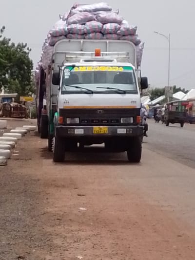 A Kia truck, heavily laden with sacks, stands on the side of a road in what appears to be Dungu, Ghana. The truck has a Ghanaian license plate. The area shows signs of commercial activity. Some people are walking.