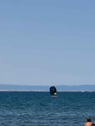 In the distance, a person is wing foiling on the Black Sea near Atiya, Bulgaria. They are wearing a yellow shirt and are on a white board, holding a blue and orange wing. The water is choppy, and a distant city skyline is visible on the horizon. In the foreground, a person is partially visible from the shoulders up, looking out at the water.