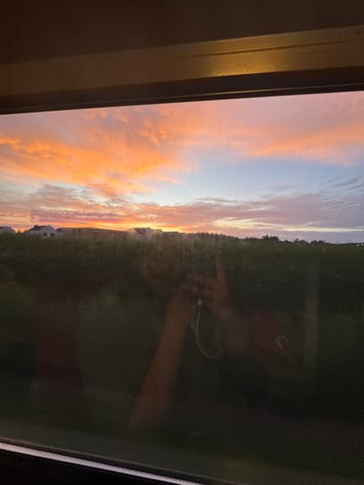 The image captures a sunset viewed through a window in Palędzie, Poland. The sky is ablaze with vibrant hues of orange, pink, and blue, with scattered clouds. In the foreground, the reflection on the window shows a person holding a smartphone and wearing earbuds. Beyond the window, a dark, green landscape is visible, with the silhouettes of buildings in the distance. The overall scene conveys a sense of a quiet evening observation.