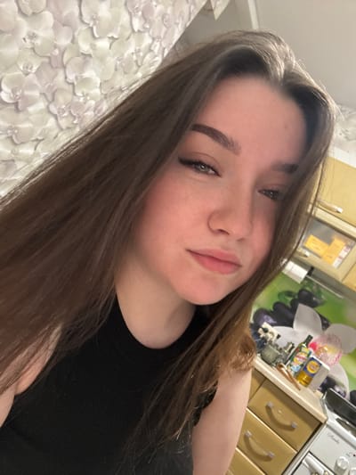 This media file is a close-up, head-and-shoulders selfie of a young woman. She has long, dark brown hair that is styled casually and falls around her shoulders and face. Her skin is fair with a rosy complexion, and she appears to be wearing minimal makeup, with a touch of blush on her cheeks and mascara on her eyelashes. Her eyes are a light green color, and she is looking directly at the camera with a neutral or slightly amused expression.
The background is somewhat blurred but suggests an indoor setting. To the left, there is a textured surface with a white, floral pattern, possibly a blanket or upholstery. To the right, kitchen cabinetry is visible, including wooden drawers with silver handles and a countertop. On the countertop, there are several small objects, including bottles and containers of various brands, one of which has text that is not clearly legible but appears to be for food or cleaning products. Above the cabinets, a section of a wall with a graphic depicting flowers and possibly fruit can be seen. The lighting is somewhat dim, suggesting it might be evening or the room is not brightly lit. The overall atmosphere is informal and personal, typical of a selfie taken at home. The location context provided is Gamovo, Russia.