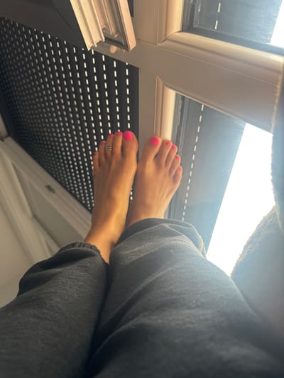 The image captures an interior scene in Nogent-sur-Marne, France, viewed from a low perspective. A pair of bare human feet, positioned centrally and slightly elevated, are the primary subject. The skin of the feet exhibits a warm tone. All visible toenails are painted a vibrant hot pink. A metallic, multi-band toe ring is present on the second toe of the left foot. The individual's lower legs are partially visible, covered by dark grey or black sweatpants, which display minor fabric pilling. In the background, a white-framed window is visible, with a partially lowered dark-colored roller shutter on the left side, featuring small, horizontal light-colored slits. The right portion of the window displays an overexposed bright white area, indicating strong ambient light from outside. The feet appear to be resting or elevated towards the window.
