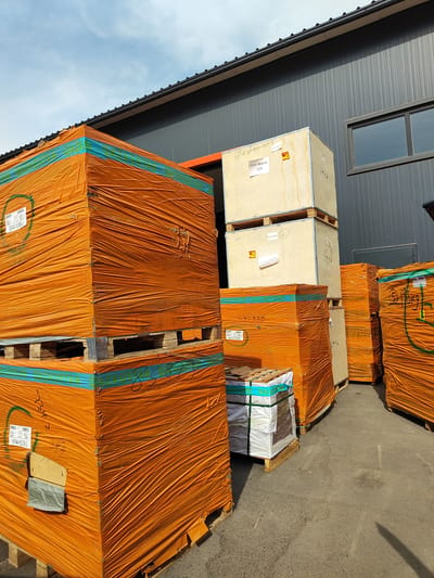 This media file depicts an outdoor storage area, likely a loading dock or warehouse exterior, in Boralday, Kazakhstan. The scene is dominated by large, stacked pallets of goods wrapped in bright orange plastic sheeting. The pallets are of varying sizes and appear to be made of wood. Some of the stacks are very tall, suggesting significant quantities of material.
In the background, a modern-looking industrial building with a dark gray, ribbed facade and a black roof is visible. A window with a reflective surface is present on the building. The sky above is partly cloudy with a light blue hue, indicating daytime.
Notable details include various labels and markings on the wrapped pallets and wooden crates. Some visible text on a light-colored wooden crate reads "ASA-Marco 1/3" and "ASA2410-06/07-3". Another crate has a label with what appears to be a barcode and more text. Some of the orange wrapping also has blue banding with handwritten numbers, such as "85873". One smaller pallet in the foreground contains items wrapped in white and clear plastic.
The overall impression is one of a commercial or industrial setting where goods are being stored or prepared for shipment. The bright orange wrapping stands out against the muted tones of the building and the asphalt ground. The lighting suggests it is a sunny day. No people are visible in the frame. The stacked nature of the goods and the presence of the industrial building strongly suggest a logistical or warehousing context.