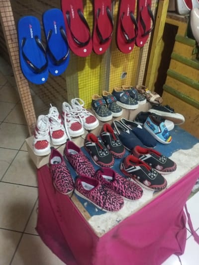 Displayed on a makeshift table draped with pink fabric is an assortment of footwear. Pairs of blue and red flip-flops hang above. Below are children's sneakers in various designs. Red and white sneakers and dark slip-ons with red or pink patterns are visible. More children's shoes are placed near the back including dark blue with spider-man and brown sandals.