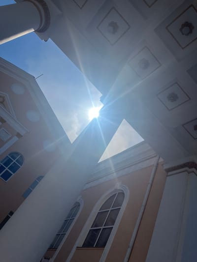 This is a low-angle, wide shot of the sun shining brightly through a gap between two buildings, creating lens flare. The buildings appear to be made of light-colored stone or stucco, with ornate architectural details. The building on the left has several arched windows. The building on the right has a patterned ceiling visible. The sky is a clear blue. The location is the Karaganda Palace of Culture in Temirtau, Kazakhstan.