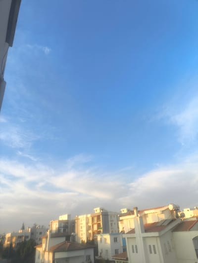 The image displays an elevated view of a residential urban area within Nicosia, Northern Cyprus / Cyprus, captured under a predominantly clear blue sky with scattered, wispy white clouds. The upper two-thirds of the frame are occupied by the sky, transitioning from a deeper blue at the top to a lighter hue near the horizon, with diffuse cloud formations. The lower third features a densely packed collection of multi-story buildings, primarily constructed with light-colored facades (white, beige, light grey). Architectural elements include flat roofs and pitched roofs with red/orange tiles. Multiple rooftops feature visible solar water heaters and photovoltaic panels. Green foliage from trees and shrubs is interspersed among the buildings, particularly on the left and center-left. A dark, vertical structural element, likely part of a building or balcony railing, frames the extreme left edge of the image. No individuals, vehicles, or discernible activities are present in the scene. The overall lighting condition suggests either early morning or late afternoon, with a warm, soft illumination on the structures.