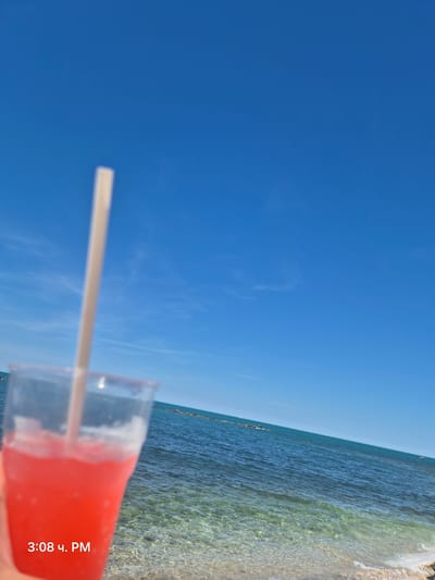 A bright, sunny day at the beach in Bisceglie, Italy. The foreground features a hand holding a clear plastic cup filled with a vibrant pink frozen beverage, likely a slushy or granita, with a light-colored straw. Beyond the drink, the crystal-clear waters of the Adriatic Sea stretch towards the horizon under a brilliant blue sky with sparse white clouds. The seabed is visible through the shallow, transparent water, revealing a mix of sand and pebbles. In the distance, a line of dark, rocky structures emerges from the water. The timestamp "3:08 Ч. PM" is superimposed on the bottom left of the image, indicating the time of day.