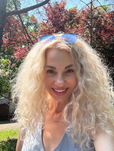 In a selfie taken outdoors, a blonde woman with curly hair and sunglasses perched on her head smiles at the camera. She is wearing a light blue top and a silver necklace. The background consists of trees with red and green leaves, and a clear blue sky. The sunlight is bright, casting shadows and highlighting the woman's features. The setting appears to be a park or garden, possibly in Goryachy Klyuch, Russia, given the vibrant foliage and sunny weather.