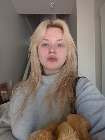 The image displays a head-and-torso view of a female individual situated indoors. The subject has shoulder-length blonde hair, light-colored eyes, and a fair complexion, facing directly towards the camera with slightly pursed lips. She is dressed in a plain light grey, high-necked, long-sleeved garment. A dark strap, possibly from a bag, is visible over her right shoulder. In the lower right foreground, a textured, light brown, furry object is partially visible. The background features plain grey walls, with a lighter grey vertical panel visible on the left and a uniformly light grey wall on the right. A light-colored ceiling with a small, circular white fixture is visible above the subject's head. There are no identifiable landmarks or environmental cues within the image to specify the location as Area D (Elaho/Garibaldi), Canada.