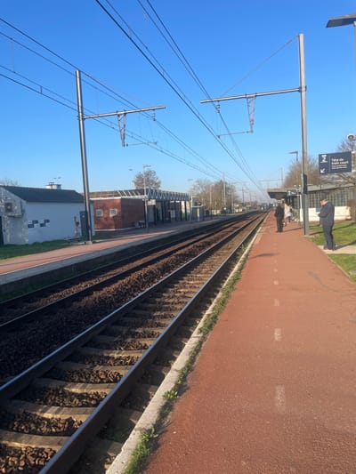 This image depicts a train station platform at Plaisir-Grignon station in Plaisir, France, under a clear blue sky. The view is along the length of two parallel train tracks, receding into the distance.

In the immediate foreground, a reddish-brown textured platform surface is visible, featuring white dashed lines. A concrete curb with sparse grass and weeds separates this platform from the tracks. Two sets of active railway tracks, composed of steel rails, wooden sleepers, and gravel ballast, run parallel from the foreground to the horizon.

On the right platform, in the mid-ground, a metal pole supports a blue informational sign with a white arrow pointing left and text indicating "2 Tête de train court" (Platform 2, Short train head) with additional smaller text below. Adjacent to the sign, a male individual in a dark jacket and trousers stands, appearing to look at a mobile device. Further down the platform, a group of three individuals, including two adults and one smaller figure, are gathered. One adult wears a dark coat, the other a light-colored coat. Overhead, a complex network of catenary wires for electric traction is suspended by multiple tall metal poles.

On the opposite side of the tracks, a second, parallel platform is visible. This platform features a small building with a reddish-brown base and a lighter-colored roof, likely a station shelter or utility building. Another smaller, white-walled building with a dark roof is situated further left on the same platform. The background reveals distant trees and faint outlines of structures, suggesting a suburban or semi-rural environment. The scene is brightly illuminated by daylight, casting shadows towards the right.