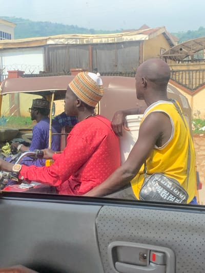 In this rotated image, three individuals are observed within or near a vehicle, possibly a tuk-tuk. The primary focus is on two men in the foreground. One man, wearing a yellow sleeveless jersey and a black messenger bag with a geometric pattern, leans forward. The other man, in a red patterned shirt and a white and orange striped cap, is also positioned similarly, with his right hand resting on a steering mechanism. Both men have dark skin and appear to be engaged in a conversation or transaction.
A third person, wearing a purple shirt, is visible in the background, partially obscured. The scene appears to be outdoors, with buildings and vegetation visible in the distance, suggesting an urban or semi-urban environment in Dam Camp, Nigeria. The lighting suggests it is daytime, and the weather appears to be clear. The overall impression is one of everyday activity, possibly related to transportation or commerce.
There is no visible text in the image. The image is a candid shot, capturing a moment in time without any posed elements. The expressions of the individuals are not clearly discernible due to the angle and resolution. The presence of the tuk-tuk suggests it is a mode of transport commonly used in the region.
