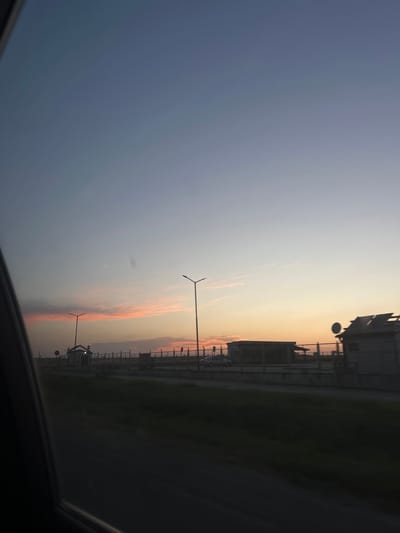 This media file depicts an outdoor scene at dusk, likely in Plodovitovo, Bulgaria. The image is oriented vertically. The foreground is dominated by the dark interior of a vehicle, suggesting the photo was taken from inside a car or bus.
The horizon is visible, separating the sky from the landscape. The sky exhibits a gradient of colors, transitioning from a deep blue at the top to soft oranges and pinks closer to the horizon, indicative of sunset. Wispy clouds are scattered across the sky, tinged with the colors of the setting sun.
In the midground, there's a fence line running horizontally across the image. Beyond the fence, several structures resembling buildings or industrial facilities are visible. Two lampposts, appearing modern and minimalist in design, stand along the fence line. In the distance, there are hints of green fields or grassy areas. The overall atmosphere suggests a somewhat rural or industrial setting on the outskirts of a town.
The time of day is clearly evening, with the low-lying sun casting a warm glow. The weather appears calm, with no strong indications of rain or wind. The image doesn't contain any visible text. The scene evokes a sense of transition, likely the end of a day, with a peaceful yet somewhat industrial feel.