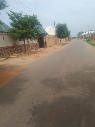 The image depicts a street scene in Gindi Nassarawa Kyarmi Galbi, Nigeria. The street is paved with asphalt and is empty except for a single person on a bicycle visible in the distance. On the left side of the street, there is a beige wall with a brick pattern and a green-roofed structure. Further down the street, more houses with similar fencing and green roofs can be seen. On the right side, there is a line of houses with beige walls and fences, and some vegetation. The sky is overcast.