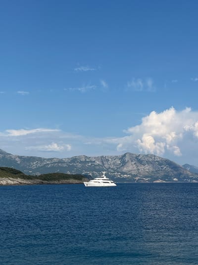 This media file depicts a serene seascape with a prominent mountain range. The scene is divided horizontally, with the upper portion dominated by a clear blue sky dotted with scattered white clouds. The lower portion features a vast expanse of deep blue water, reflecting the sky and creating a textured surface with gentle ripples.
Emerging from the water and hugging the base of the mountains is a steep, verdant slope, suggesting a rugged coastline. The mountains themselves are rugged and rocky, with patches of lighter terrain visible, possibly indicating scree or snow. A modern white vessel, likely a yacht or small cruise ship, is visible navigating the water close to the shore, its sleek form contrasting with the natural landscape.
The overall impression is one of a bright, sunny day, likely during daylight hours, given the clear sky and vibrant colors. The location appears to be a coastal area with significant elevation changes, characteristic of a fjord or a mountainous coastline. There is no visible text in the image. The atmosphere conveyed is peaceful and majestic, highlighting the grandeur of nature.
