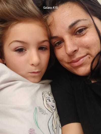 A close-up, top-down shot captures two individuals, appearing to be a mother and daughter, lying down closely together. Both subjects' heads are tilted towards the right, looking towards the camera with soft, calm expressions, suggesting an intimate selfie or portrait.
The younger individual, a child, is positioned in the upper-left part of the frame. She has light brown hair and brown eyes, and her face is smooth. She is wearing a light-colored garment, possibly white or light pink, featuring a partial illustration of Elsa from Disney's "Frozen" along with subtle purple and green floral or leafy designs on the fabric.
The adult woman occupies the lower-right portion of the image. She has darker brown hair and brown eyes, with a slight, gentle smile on her face, conveying warmth and contentment. Her skin shows a few freckles and a mole on her chin. She is dressed in a dark, likely black, top or sweater.
The overall tone of the image is affectionate and peaceful, highlighting the close bond between the two. The lighting is even and natural, consistent with an indoor setting. Along the right edge of the image, the text "Galaxy A15" is visible vertically, indicating the camera used. The location context provided for the image is Obrochishte, Bulgaria.