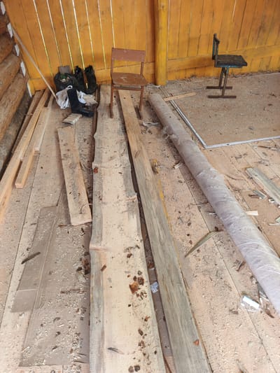 The image depicts the interior of a wooden structure, likely a shed or workshop, in the village of Islambaevo, Russia. The floor is covered in sawdust and wood scraps, indicating construction or renovation work. Several long wooden planks are laid out on the floor. In the background, a yellow-painted wooden wall serves as the backdrop. Two chairs are visible: one is a simple wooden stool, and the other is a metal chair with a worn seat. A black plastic bag is piled with what appears to be black boots. A large roll of material, possibly protective covering or insulation, is also present. The overall scene suggests a utilitarian and unfinished space.