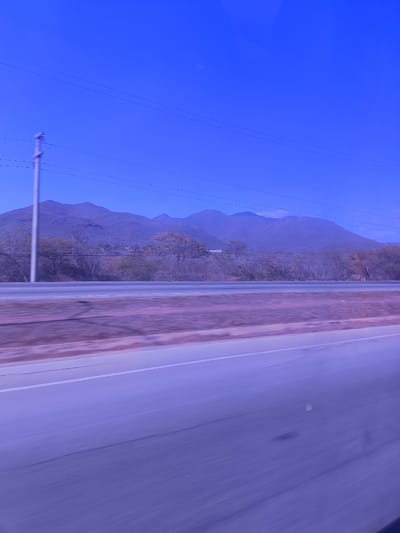 A daytime photograph captures a roadside view in what appears to be El Dátil, Venezuela. The sky is clear blue. A mountain range is visible in the background. In the foreground, a highway stretches into the distance, marked by a white line. An utility pole is visible on the left of the picture. The photograph seems to be taken from a moving vehicle. Trees line the roadside. There are no visible people or interactions.