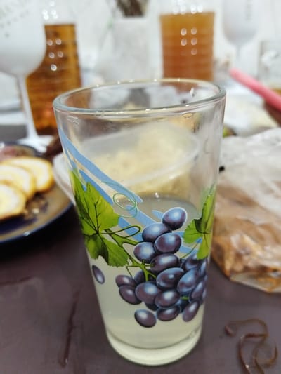 A close-up shot focuses on a clear glass with a grape vine design, filled with a pale yellow liquid. The glass sits on a dark, reflective surface, possibly a table, which has a small spill of liquid on it. In the background, out of focus, are other glasses, one containing amber-colored liquid, and what appears to be a white ceramic container. To the left of the glass, on a dark blue plate, are several slices of a rolled cake or pastry with a light yellow filling. The scene is indoors, suggesting a domestic or dining setting in Novostroika, Russia.