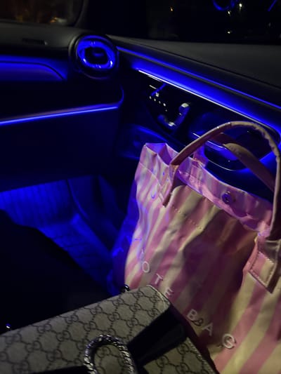 The image captures the interior of a vehicle at night, dominated by a vibrant blue ambient lighting scheme. On the right side, a pink and white striped tote bag is prominently displayed, with the words "TOTE BAG" visible on its surface. Below the tote bag, a patterned strap, featuring repeating "GG" logos often associated with the Gucci brand, is partially visible. The car's dashboard or door panel is illuminated by a long, thin strip of blue light, creating a futuristic ambiance. A circular air vent on the left side of the frame also glows with blue light. The deep blue light reflects on the metallic and plastic surfaces, highlighting contours and details within the car. The darkness outside the car windows confirms it is nighttime. No people or specific activities are depicted; the focus is on the illuminated car interior and the personal items within it.