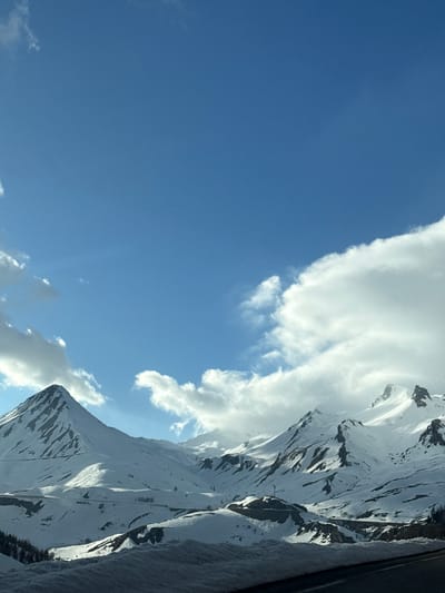 The image shows a view of snow-covered mountains under a partly cloudy blue sky. The mountain range dominates the right side of the frame, with the snow highlighted by sunlight. Clouds are interspersed among the peaks and in the sky. The location is Le Monêtier-les-Bains, France.