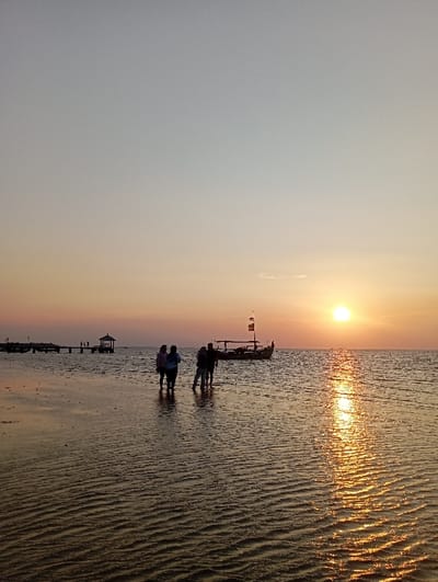 This image depicts a coastal scene at sunset in Tritunggal, Indonesia. The sky transitions from a pale grey-blue overhead to warm orange and pink hues at the western horizon. A bright, circular orange sun is positioned just above the horizon line, casting a prominent, shimmering golden reflection path across the calm, shallow ocean water. The water in the foreground and midground covers a textured seabed, exhibiting distinct ripple patterns.
Five human figures are present in the shallow water, partially silhouetted. Three individuals are grouped on the left side of the frame, facing towards the horizon. Two additional individuals stand further to the right, closer to a traditional-style boat. This boat, dark-hulled with multiple vertical poles and flag-like adornments, is anchored or beached in the shallow water towards the right-center of the frame. In the far left background, two wooden pier structures extend into the water; one is a longer pier with multiple posts, and the other is a shorter pier terminating in a small, covered gazebo-like structure. The individuals appear to be observing the sunset or the surrounding coastal environment.