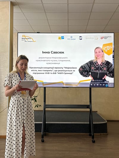 This media file captures a presentation taking place indoors, likely in a community hall or conference room. The primary subject is a woman, identified by the text on the screen as Inna Savsyuk, the director of the Myronivka Regional Museum. She is standing in front of a large screen displaying presentation slides, and she is holding a document and appears to be speaking.
The screen prominently features text in Ukrainian, indicating the presentation is about a project titled "Myronivka: The City That Speaks." It also mentions that the project is being implemented with the support of the Ukrainian Cultural Foundation and the MHP-Community Foundation. The screen also displays logos of supporting organizations, including "Myronivka City Council" and the "Myronivka Regional Museum." A photograph of Inna Savsyuk is visible on the top right of the screen.
Inna Savsyuk is wearing a black and white polka dot dress and a lanyard with an identification badge. The overall atmosphere appears professional and informative, with the woman engaged in delivering her presentation. The lighting suggests it is daytime, and the indoor setting implies no specific weather conditions are relevant. The scene is set in Myronivka, Ukraine, as indicated by the text on the screen.