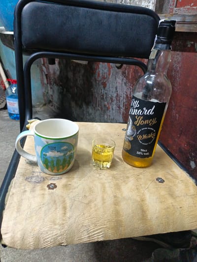 On a rough, weathered wooden surface, likely a table or chair seat, are three objects: a mug, a shot glass, and a bottle. The mug, decorated with a sky and landscape scene, sits to the left. To its right is a small, clear shot glass containing a yellow liquid. To the right of the shot glass is a partially empty bottle of "Bernard Honey" spirit, with a label indicating it is 70cl and 35% vol. The bottle has a black cap with peeling seals. The background consists of a dark, textured surface, possibly a wall or furniture, with hints of blue and red paint. A black metal chair frame is visible supporting the wooden surface. The scene appears to be outdoors or in an unrefined indoor space, with the image capturing a moment of implied consumption or preparation for it. The location is identified as Pivdenne, Ukraine.
