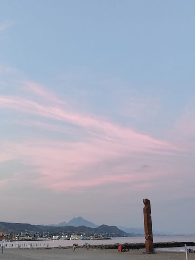 The image captures a sunset scene at the beach in El Campello, Spain, with a tall, abstract, metallic sculpture dominating the right side of the frame. The sky is a gradient of soft pinks and blues, with wispy clouds stretching across it. In the background, a coastline with buildings, a harbor with boats, and rolling hills or mountains are visible. The foreground shows a sandy beach with a few people scattered around, some seated under umbrellas. The sculpture, made of what appears to be weathered metal, has a vertical orientation with angular cutouts.