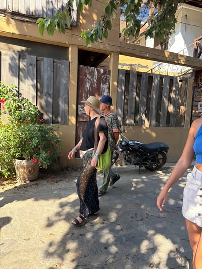 The image depicts a sunlit street scene in Mazunte, Mexico, featuring three individuals and a parked motorcycle amidst local architecture.

In the foreground, on the right, the right arm, shoulder, and upper torso of an individual are visible, wearing a blue crop top and white, button-front denim shorts. This individual's right hand is slightly open.

In the midground, to the left, a fair-skinned individual with visible gray hair wears a light-colored straw hat, a black t-shirt, and dark wide-leg pants with a light-colored, possibly peacock feather, pattern. This person has a light blue wristband and a blue-faced watch on the left wrist, carries a small light-colored fanny pack/pouch on the left hip, and a bright green shoulder bag over the left shoulder. They are walking towards the right, wearing dark sandals.

Immediately behind this individual, slightly to the right, another fair-skinned individual walks in the same direction. This person wears a dark blue bucket hat, a patterned long-sleeve shirt (possibly black and white), and light-colored pants with dark shoes.

A dark-colored motorcycle, possibly black, is parked on the textured, stone-paved ground to the right of the walking individuals. Overhead, tree branches with green leaves cast shadows across the ground and buildings.

The background consists of a building with light brown/tan stucco or adobe walls. The lower level features wooden shutters on a window and a weathered, dark brown wooden door. Above this, a lighter tan section is visible with what appears to be part of a balcony or upper story with railings. To the right, another section of a building facade shows vertical wooden slats and a partially visible white "E" sign, likely indicating "Estacionamiento" (Parking). Exposed wires run horizontally across the scene at various heights.

On the far left, a large terracotta pot holds a vibrant green plant with numerous red flowers. The ground is an uneven surface of stone slabs, with strong sunlight illuminating parts of it and distinct shadows from the overhead foliage.