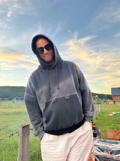 A man wearing sunglasses and a grey hoodie is standing outside in a grassy field with a barbed wire fence in the foreground. In the background, there are some wooden buildings, hills with trees, and a cloudy sky with hints of yellow and orange from what appears to be a sunset. The man has his hands in his pockets and is smiling at the camera. To the right of the man, there is a fire pit with a pot on it, and two containers covered in plastic wrap. This scene is located in Virak, Montenegro.