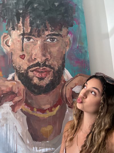 A woman is posing in front of a large painting of a man. The painting is abstract and features bold brushstrokes and vibrant colors, with a teal and pink background. The man in the painting has dark, curly hair, a beard, and is wearing a white shirt with a red and yellow beaded necklace. He has a red heart shape painted on his cheek and a yellow heart shape on his chest. The woman, who has long, wavy brown hair with braids, is looking up and to the side with her lips pursed, as if about to kiss. She is wearing sunglasses on top of her head and a necklace. The painting appears to be quite large, taking up a significant portion of the background. The image is shot from a slightly low angle. The overall scene suggests a personal or artistic setting, possibly a home or studio. The question of location in San Juan, United States of America is not supported by any visual evidence in the image itself.