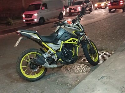 A yellow and black motorcycle is parked on the side of a street near a manhole cover and a storm drain. The street is paved with asphalt. Several vehicles are visible in the background, including a white van, a silver SUV, a yellow taxi, and an orange car. The scene appears to be at night, with streetlights providing illumination.