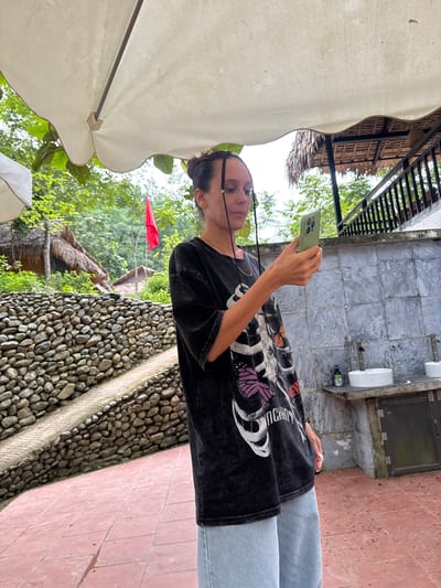 In Cổ Lũng Commune, Vietnam, a woman stands outdoors under a large white umbrella, holding a light green smartphone in her right hand and looking at its screen. She wears a dark t-shirt with a skeleton and butterfly graphic and light blue wide-leg pants. The background features lush green trees and vegetation. To the left, a textured stone wall with a paved pathway runs diagonally. To the right, a concrete structure with sinks and faucets is visible, along with dark railing and a thatched-roof building. A red flag is planted in the distance. The ground is paved with reddish-brown tiles.