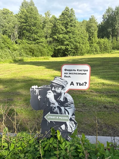 The image depicts an outdoor scene in Sestroretsk, Russia, featuring a cutout of a person holding a vintage film camera. This cutout is placed in a grassy area with trees in the background, suggesting a park or natural setting. The weather appears to be sunny with some clouds.
Two signs are prominently displayed next to the cutout. The first sign, with a red border, contains text in Russian: "Фидель Кастро на экспозиции был." which translates to "Fidel Castro was at the exhibition." The second sign, a speech bubble pointing to the cutout, also in Russian, reads: "А ты?" which means "And you?". Below the cutout, another sign with an arrow states "ОТВЕТ В МУЗЕЕ" which translates to "ANSWER IN THE MUSEUM".
The overall impression is that of an interactive exhibit, possibly designed to engage visitors by posing a question and directing them to a museum for the answer. The cutout and signs are likely part of an open-air display. The time of day seems to be daytime, given the lighting. The surrounding greenery indicates it is likely spring, summer, or early autumn.