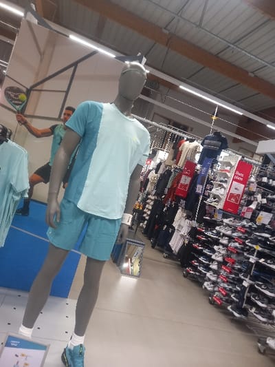 The image displays the interior of a sporting goods retail store in Vendin-le-Vieil, France.
In the foreground, a grey, headless mannequin stands centrally, facing slightly right. It is attired in a light blue and white color-blocked short-sleeved athletic shirt, light blue athletic shorts, white athletic socks, and light blue athletic shoes accented with yellow. A white wristband is present on its right wrist. A partial product display stand is visible near the mannequin's left foot, and a small, illuminated rectangular display is positioned on the floor behind its lower legs.
In the mid-ground to the left, a male individual with dark hair wears a teal athletic t-shirt and dark shorts. He is captured in a dynamic pose, holding a green and black padel racket with a yellow ball on its face, extended upwards with his right arm, simulating a serve or overhead shot. He stands on a blue mat marked with white lines, indicative of a court section.
The background on the right side of the frame features extensive retail shelving and racks densely stocked with athletic apparel and footwear. Numerous rows of athletic shoes, primarily in white, black, and dark blue, are displayed on shelves. Clothing items hang on racks. Red signs bearing white text, one partially visible as "Bons plans," are interspersed among the merchandise. The store's environment includes a light-colored floor, a high ceiling with exposed wooden beams and metal roofing, and fluorescent strip lighting fixtures. Additional merchandise displays are present in the deeper background to the left.