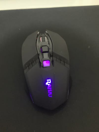 A black computer mouse rests on a dark mousepad. The mouse features a glossy black finish with geometric details and an illuminated "RAIKU" logo in purple. A single button near the front of the mouse is also illuminated in purple. The mousepad appears to be a smooth, matte black surface. There is nothing in the image to indicate a location in Ijeododo, Nigeria, and the scene appears to be indoors, possibly on a desk or similar surface.