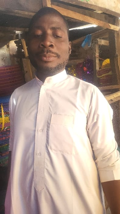An adult male is depicted in a medium close-up, standing in an indoor or semi-sheltered commercial area in Fagge D1, Nigeria. The subject, of African descent, wears a long-sleeved white garment with a band collar featuring two small button-like embellishments, a buttoned placket, and a left chest patch pocket. He has a dark beard and mustache and short dark hair, and is looking directly forward. The background on the left shows stacks of vibrantly colored woven items, likely mats or rugs, displaying patterns in red, blue, green, yellow, and purple. To the right, a wooden frame structure is visible, partially obscuring mesh screening behind which various colorful, reflective, or plastic objects are present, including yellow fan-shaped items. Exposed wooden support beams are visible overhead.