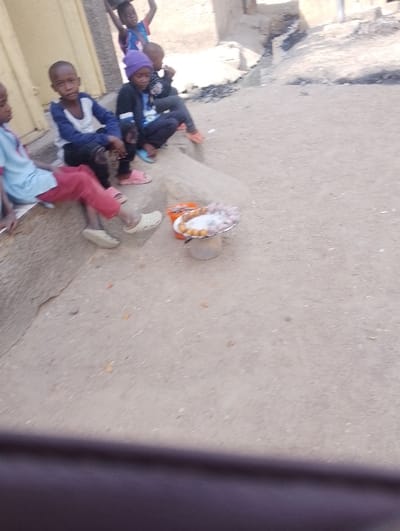 The image depicts an outdoor scene in Dorayi, Nigeria, featuring six children and a makeshift food stall on an unpaved earthen ground. In the foreground, four children are seated on a low concrete step or ledge. To the far left, partially out of frame, a child wears a light blue shirt and red pants. Next to them, a child in a blue and white long-sleeved shirt and dark pants faces forward. Further right, a child wearing a purple beanie, dark jacket, and dark pants sits lower on the step, looking towards the ground. Another child sits behind them, wearing a dark top and orange footwear.
In front of the seated children, a small, elevated display holds an assortment of round, fried food items on a metallic plate, supported by a cylindrical base. Adjacent to this display are two orange plastic buckets, one containing more fried food items and the other holding light-colored, possibly chopped, vegetables.
In the mid-ground, slightly behind the seated children, two more children are visible. One is standing, carrying an object on their head, and wears a blue and red top. The other child is partially obscured behind them. The background features simple, light-colored buildings and an unpaved pathway with a narrow drainage ditch. The ground is dusty and light brown. The overall scene is brightly lit by natural daylight.