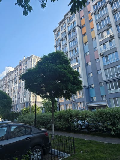 This media file depicts an outdoor scene in Petropavlivska Borshchahivka, Ukraine, likely during the daytime. The primary subjects are modern residential buildings with multiple stories, characterized by their gray facades accented with colorful panels in shades of orange, pink, and blue. The buildings feature numerous windows, and some have air conditioning units mounted externally.
In the foreground, a lush green tree with a full canopy stands prominently, partially obscuring one of the buildings. Adjacent to the buildings and tree, there is a paved walkway bordered by a strip of well-maintained green grass. A dark-colored car is parked near the walkway, partially visible behind a black metal fence. The sky above is a clear, bright blue with a few wispy clouds.
The scene appears peaceful and serene, suggesting a typical day in a residential area. The presence of air conditioning units and the vibrant greenery hint at a warm season, likely late spring or summer. The overall impression is one of a modern, well-kept urban or suburban environment. No visible text is present in the media file.