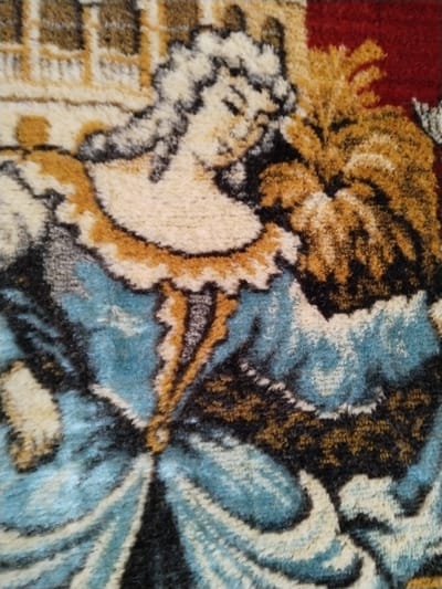 A close-up photographic detail of a high-pile textile artwork, located in Sucina, Spain, depicts an individual in period attire. The figure, possibly female, is shown from the chest up, with their head tilted downwards and to the right, featuring elaborate white or cream-colored hair or a wig and a serene expression with closed or downcast eyes. They wear a light cream bodice with a ruffled gold and brown collar, transitioning into a vibrant blue and black gown with stylized folds. The right arm holds a large, golden-brown, possibly feathered or leafy object against a dark red background. In the upper left background of the artwork, a light-colored architectural element with brown details is visible. The image showcases the coarse, tufted texture of the fabric, characteristic of a tapestry or rug.