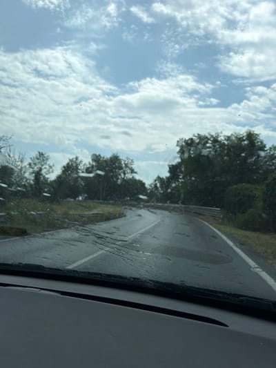 The image shows a view from inside a vehicle looking out through the windshield. The windshield is wet with water droplets and streaks, indicating rain or recent rain. The road ahead is wet and appears to be a two-lane road curving to the right. There are puddles of water on the road. The surrounding environment is lush and green, with dense trees lining the sides of the road. The sky is cloudy with patches of blue. The context of Novara, Italy, suggests this is likely a rural or suburban road outside the city center. There are no people or other vehicles clearly visible in the image. The scene depicts a drive on a wet road on a cloudy day.